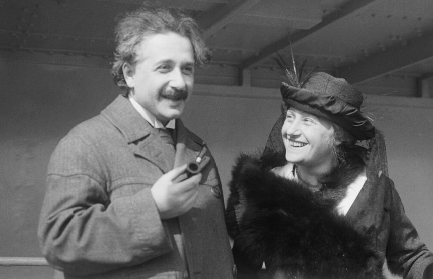 Na zdjęciu Albert Einstein z uśmiechniętą kobietą, oboje w eleganckich strojach, wesoło rozmawiają.