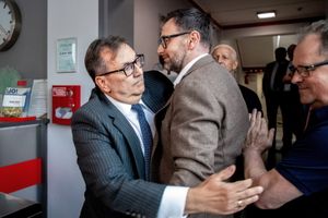 Mężczyźni w garniturach stoją blisko siebie, z widocznymi emocjami, w tle inni ludzie.