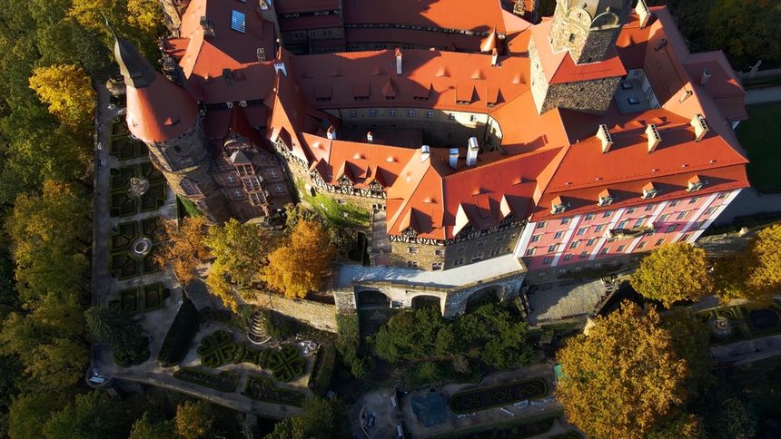 Widok z lotu ptaka na zamek otoczony kolorowym lasem, z czerwonymi dachami i ogrodami.