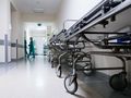 Korytarz szpitalny z metalowymi łóżkami na kółkach i personelem medycznym w zielonych strojach.