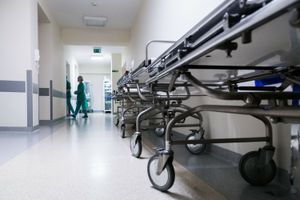 Korytarz szpitalny z metalowymi łóżkami na kółkach i personelem medycznym w zielonych strojach.