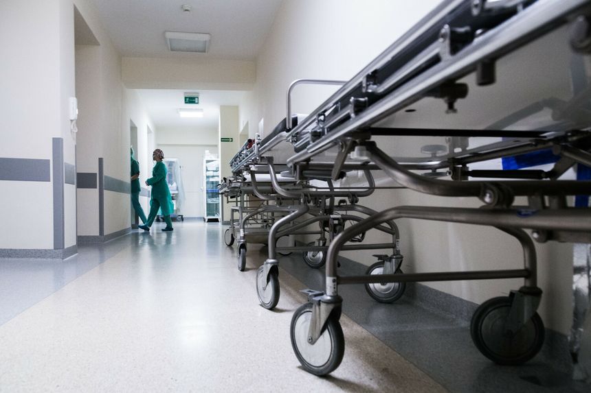 Korytarz szpitalny z metalowymi łóżkami na kółkach i personelem medycznym w zielonych strojach.
