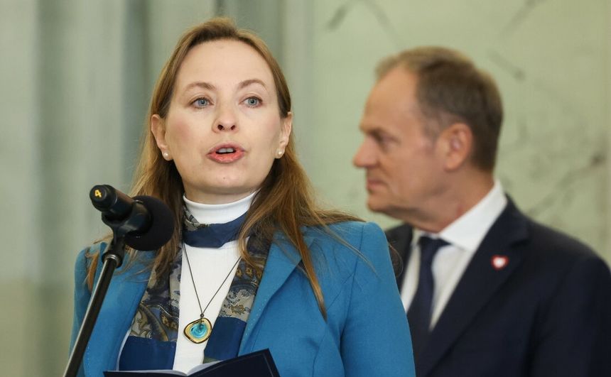 Kobieta z mikrofonem w ręku, w tle mężczyzna w garniturze. Rozmowa w poważnej atmosferze.