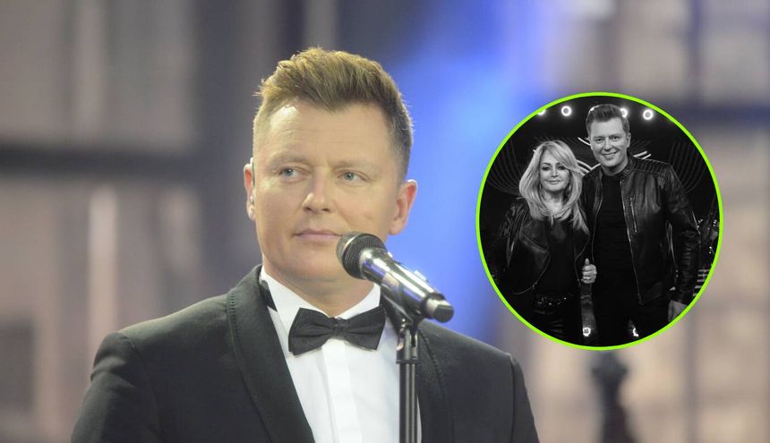 Rafał Brzozowski w garniturze z mikrofonem, na tle sceny.