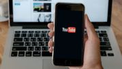 Na zdjęciu widoczny smartfon z logotypem YouTube na ekranie, w tle laptop i kawa.