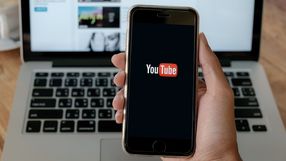 Na zdjęciu widoczny smartfon z logotypem YouTube na ekranie, w tle laptop i kawa.