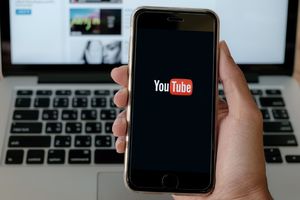 Na zdjęciu widoczny smartfon z logotypem YouTube na ekranie, w tle laptop i kawa.