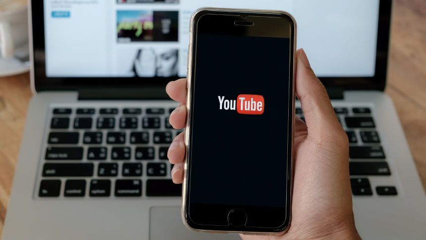 Na zdjęciu widoczny smartfon z logotypem YouTube na ekranie, w tle laptop i kawa.