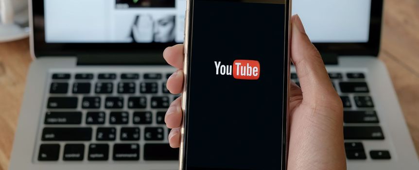 Na zdjęciu widoczny smartfon z logotypem YouTube na ekranie, w tle laptop i kawa.