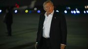 Mężczyzna w ciemnym garniturze idzie po płycie lotniska w nocy, w tle światła samolotów.