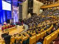Uroczystość w sali z podzieloną widownią, na scenie mówca i siedzący w toga znakujący ważne wydarzenie.