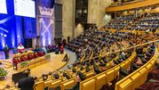 Uroczystość w sali z podzieloną widownią, na scenie mówca i siedzący w toga znakujący ważne wydarzenie.