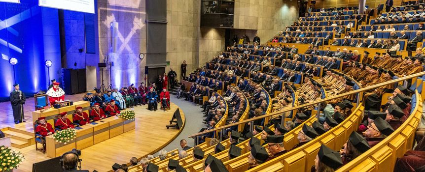 Uroczystość w sali z podzieloną widownią, na scenie mówca i siedzący w toga znakujący ważne wydarzenie.
