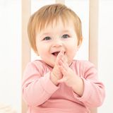 Uśmiechnięte dziecko w różowym body klaszcze w dłonie, siedząc w łóżeczku. W tle jasne deski.