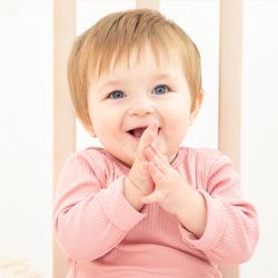 Uśmiechnięte dziecko w różowym body klaszcze w dłonie, siedząc w łóżeczku. W tle jasne deski.