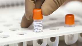 Pracownik laboratoriów trzyma próbkę w fiolce z pomarańczową nakrętką.