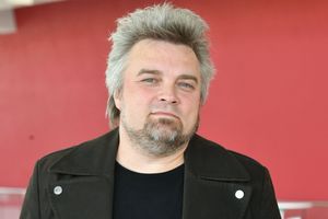 Mężczyzna z siwymi włosami w czarnej kurtce, na tle czerwonej ściany. Nie znam jego tożsamości.
