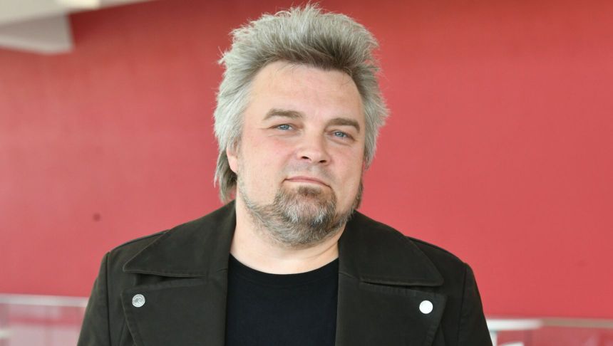 Mężczyzna z siwymi włosami w czarnej kurtce, na tle czerwonej ściany. Nie znam jego tożsamości.