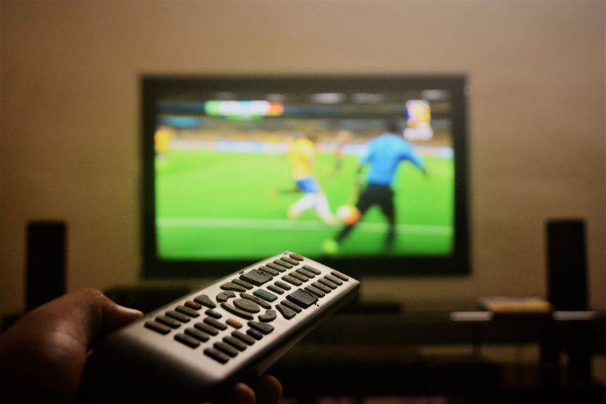 Pilota do telewizora trzyma ręka, w tle mecz piłki nożnej na ekranie telewizora.