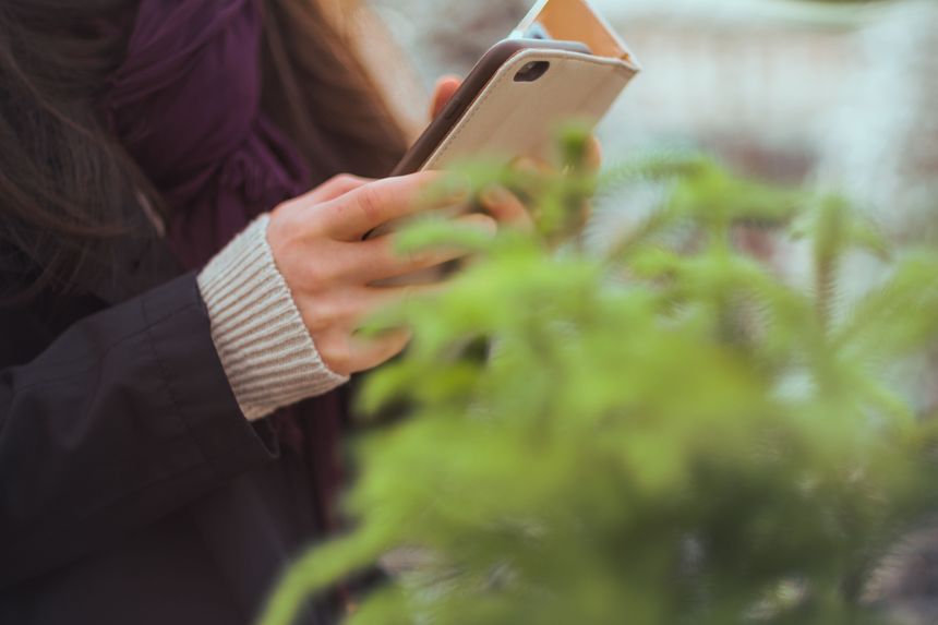 Kobieta trzyma w dłoniach telefon komórkowy, w tle widoczne liście roślin.
