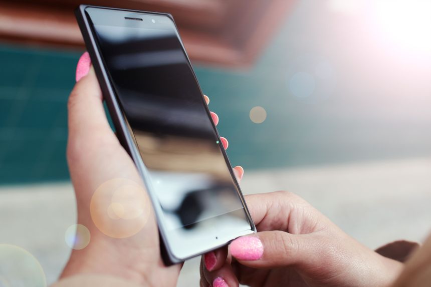 Zdjęcie dłoni trzymającej smartfon, w tle widoczne rozmyte kolory i jasne refleksy.