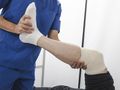 Osoba w niebieskim uniformie medycznym trzyma uniesioną nogę pacjenta z bandażem na kolanie.