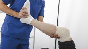 Osoba w niebieskim uniformie medycznym trzyma uniesioną nogę pacjenta z bandażem na kolanie.