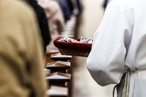 Osoba w białym stroju niesie koszyk z pieniędzmi w kościele. W tle wierni w ławkach.