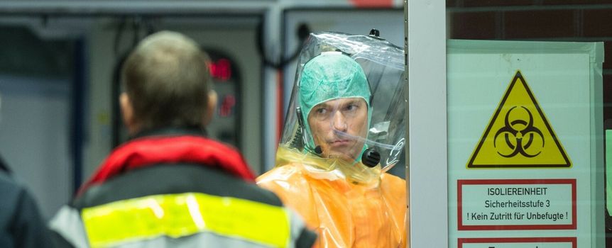 Na zdjęciu mężczyzna w pomarańczowym kombinezonie ochronnym, z maską i hełmem. W tle ostrzeżenia.