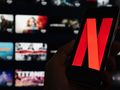 Na zdjęciu telefon trzymany w dłoni z logo Netflix na ekranie, w tle widoczne miniatury filmów.