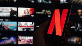 Na zdjęciu telefon trzymany w dłoni z logo Netflix na ekranie, w tle widoczne miniatury filmów.