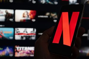Na zdjęciu telefon trzymany w dłoni z logo Netflix na ekranie, w tle widoczne miniatury filmów.
