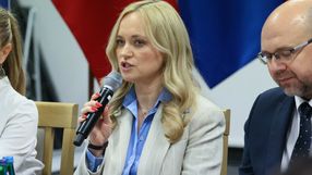 Kobieta z mikrofonem, w jasnym garniturze, dyskutuje na spotkaniu, w tle flagi.