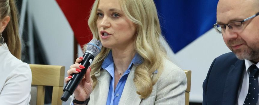 Kobieta z mikrofonem, w jasnym garniturze, dyskutuje na spotkaniu, w tle flagi.