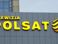 Logo Telewizji Polsat na szarej ścianie budynku, w kolorze żółtym i pomarańczowym.