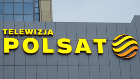 Logo Telewizji Polsat na szarej ścianie budynku, w kolorze żółtym i pomarańczowym.