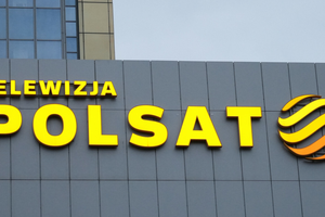 Logo Telewizji Polsat na szarej ścianie budynku, w kolorze żółtym i pomarańczowym.