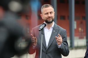 Mężczyzna w szarym garniturze przy mikrofonie, tło z budynkami.