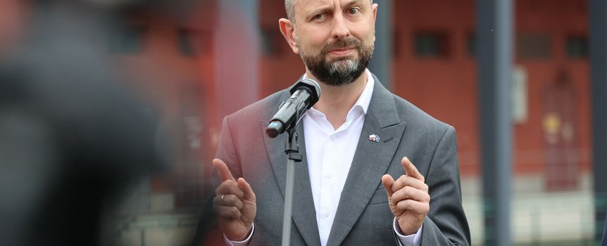 Mężczyzna w szarym garniturze przy mikrofonie, tło z budynkami.