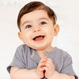 Uśmiechnięty mały chłopiec w szarym ubranku, z radosnym wyrazem twarzy, trzyma ręce złożone.