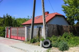 Dom z czerwoną dachówką, ogrodzeniem oraz roślinnością wokół, na tle nieba.