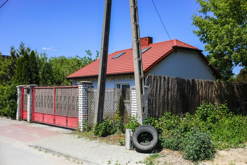 Dom z czerwoną dachówką, ogrodzeniem oraz roślinnością wokół, na tle nieba.