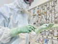 Osoba w białym kombinezonie ochronnym trzyma strzykawkę w laboratorium. W tle półki z materiałami.