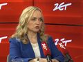 Kobieta w niebieskiej marynarce siedzi przy stole, mówi do mikrofonów. Tło czerwone z logo 24.
