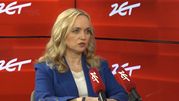 Kobieta w niebieskiej marynarce siedzi przy stole, mówi do mikrofonów. Tło czerwone z logo 24.