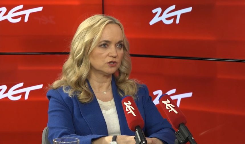 Kobieta w niebieskiej marynarce siedzi przy stole, mówi do mikrofonów. Tło czerwone z logo 24.