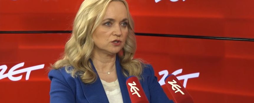 Kobieta w niebieskiej marynarce siedzi przy stole, mówi do mikrofonów. Tło czerwone z logo 24.