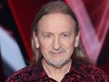 Mężczyzna z długimi włosami w kwiecistej koszuli, uśmiechający się do kamery.