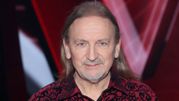 Mężczyzna z długimi włosami w kwiecistej koszuli, uśmiechający się do kamery.