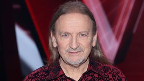 Mężczyzna z długimi włosami w kwiecistej koszuli, uśmiechający się do kamery.
